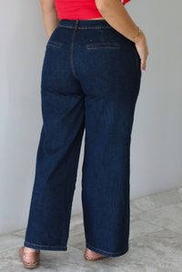 A woman shown from behind wearing wide-leg dark denim jeans, a red top, and brown woven sandals, standing indoors against a white wall with white marble flooring. 