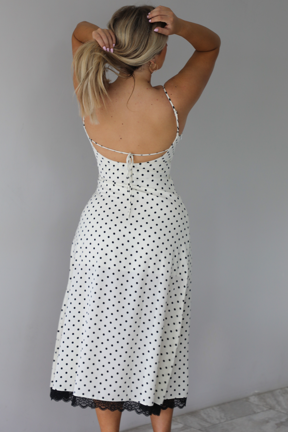 Woman wearing a white midi length dress with a black polka dot print pattern and black lace detailing at the v-neckline and bottom hem. The photo is taken indoors against a white wall with white marble flooring and shows the back of the dress, which has an open design with a tie detail in the center. 