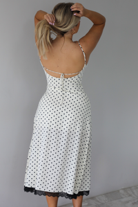 Woman wearing a white midi length dress with a black polka dot print pattern and black lace detailing at the v-neckline and bottom hem. The photo is taken indoors against a white wall with white marble flooring and shows the back of the dress, which has an open design with a tie detail in the center. 