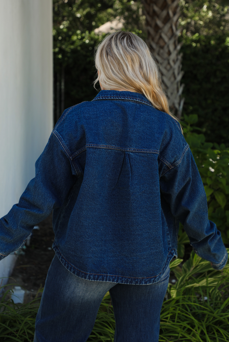 Close up of the back of a woman model wearing a dark denim button up jacket  and dark denim jeans outdoors. 