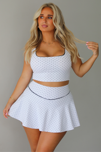 A woman modeling a white two-piece lounge set with small black polka dots, featuring a scoop-neck crop top and a matching flared skirt, standing indoors against a white wall.