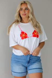 Woman wearing a white cropped boxy tee with a pink, red, and yellow bikini graphic on the front with a pair of medium wash denim shorts indoors against a white wall.