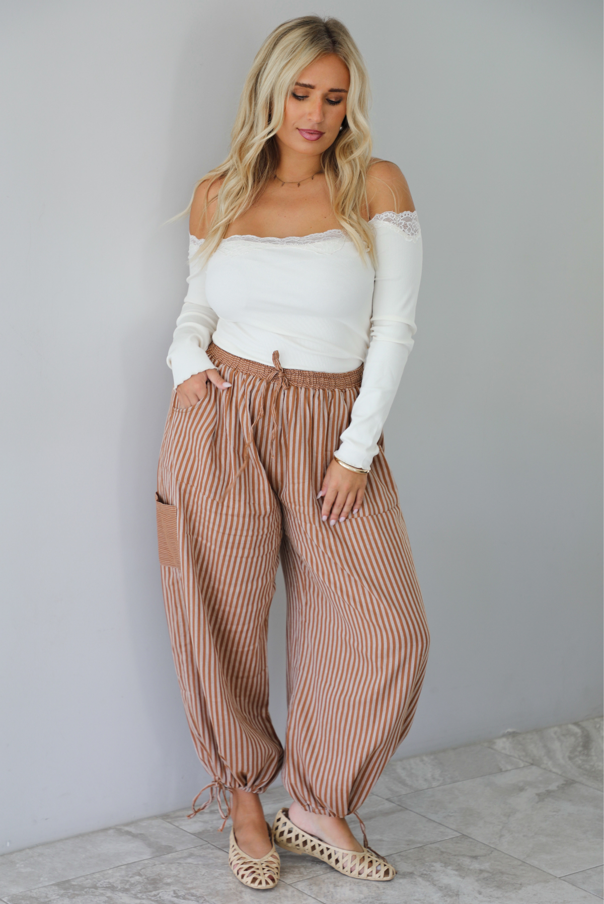 A woman wearing a cream off the shoulder long sleeved top tucked into a pair of camel and cream striped pants, standing indoors against a white wall with white marble flooring. 
