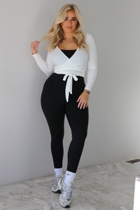 Woman wearing a white cropped wrap style fitted top over a black fitted jumpsuit. The outfit is paired with white ankle socks and silver sneakers. The photo is taken indoors against a white wall with white marble flooring. 