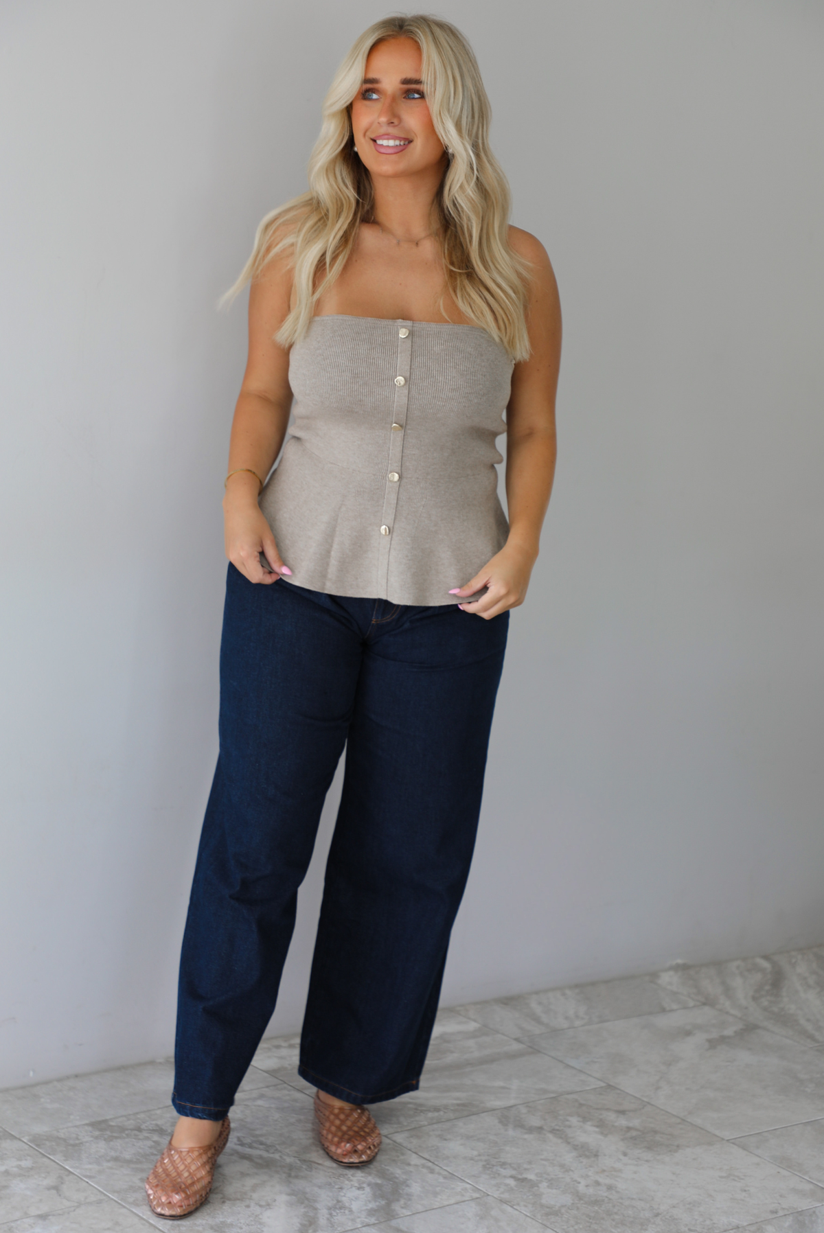 A woman wearing a beige strapless button-front peplum top and dark denim wide-leg jeans, standing indoors against a white wall with white marble flooring. 