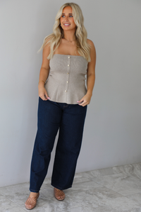 A woman wearing a beige strapless button-front peplum top and dark denim wide-leg jeans, standing indoors against a white wall with white marble flooring. 