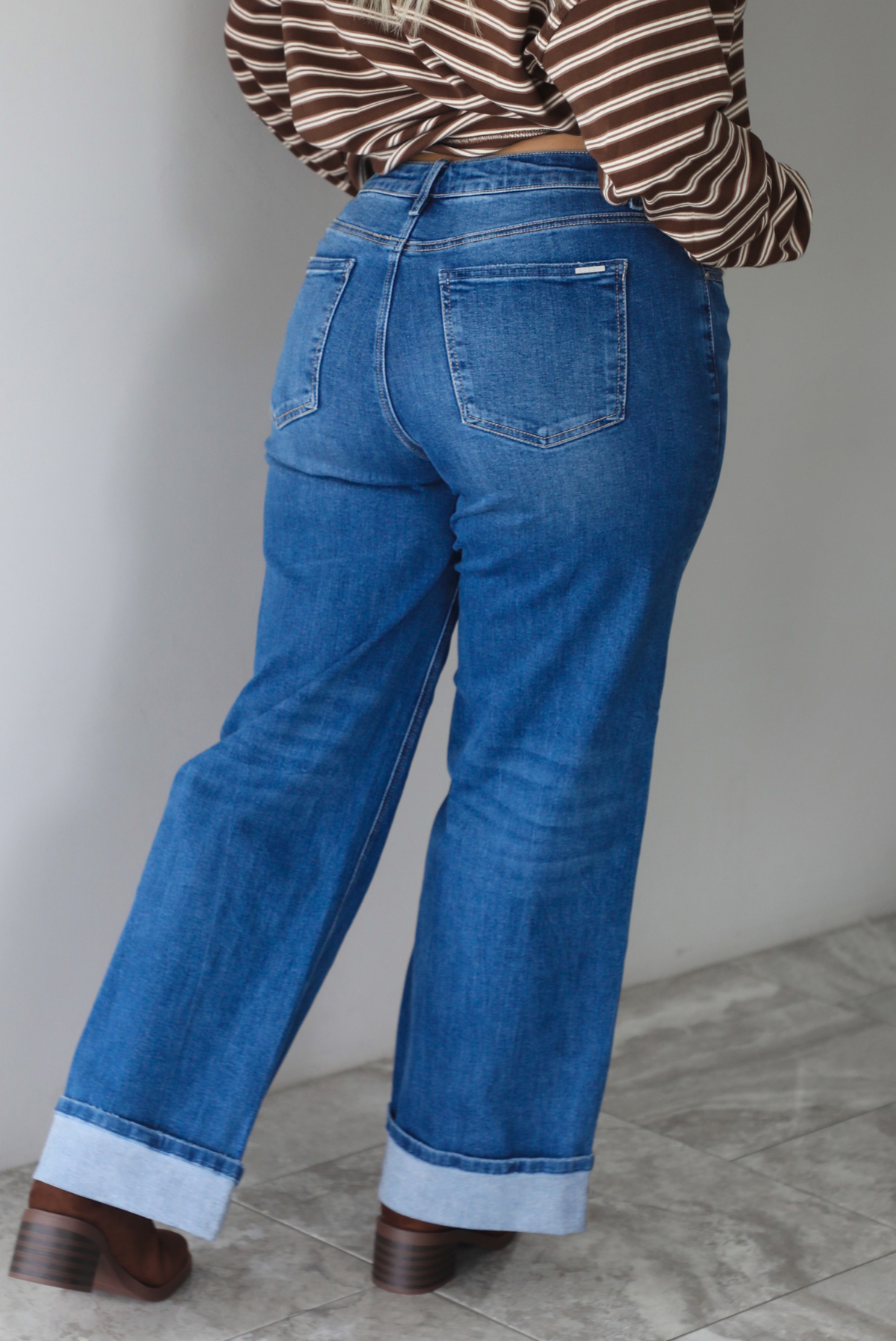 A closeup of a woman from behind wearing dark wash, wide leg denim with a cuffed hem design, standing indoors against a white wall with white marble flooring. 
