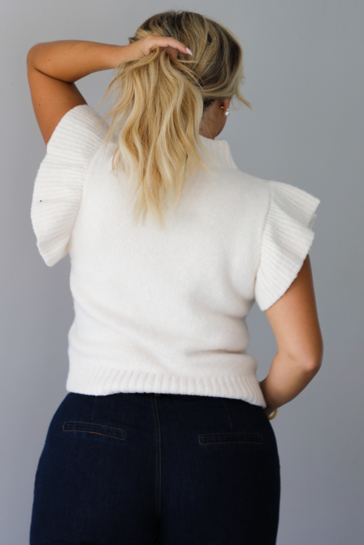 A woman shown from behind wearing a cream ribbed knit sweater top with ruffled sleeves, dark wide-leg denim jeans, standing indoors against a white wall.