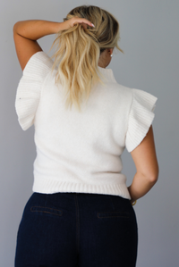 A woman shown from behind wearing a cream ribbed knit sweater top with ruffled sleeves, dark wide-leg denim jeans, standing indoors against a white wall.