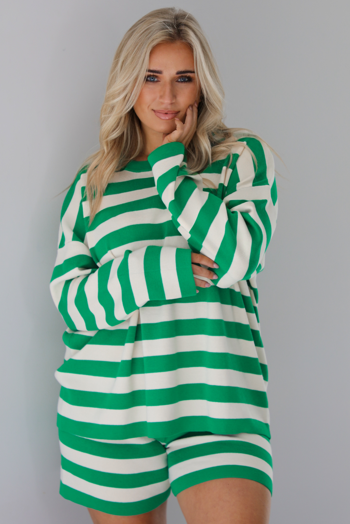 Woman wearing a green and white striped set featuring an oversized long sleeved top and high waisted shorts. The photo is taken indoors against a white wall.
