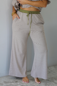 A person wearing loose, wide-leg striped pants with a muted green drawstring waistband and a matching striped crop top, standing barefoot against a plain white wall with white marble flooring. 
