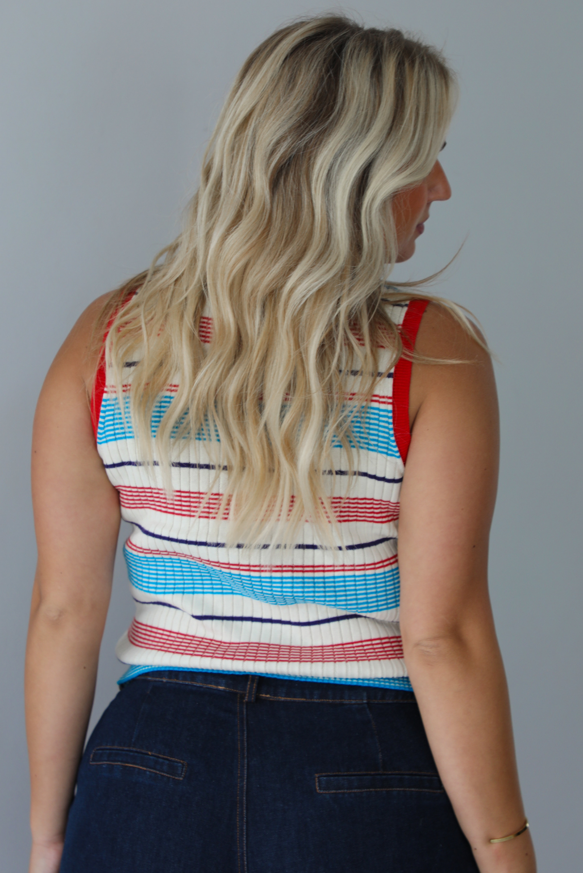 A woman modeling a sleeveless ribbed knit top with horizontal stripes in red, light blue, navy, and white, paired with dark wide-leg jeans and metallic rose gold flats, standing indoors against a white wall. The photo shows the back of the top.