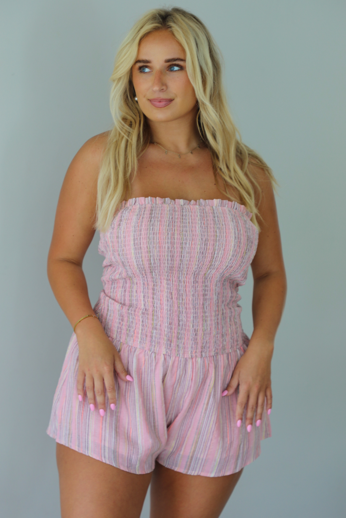 A woman wearing a pink and multi-colored vertically striped strapless romper, standing indoors against a white wall. The photo shows a closeup of the romper, displaying the smocked bodice. 