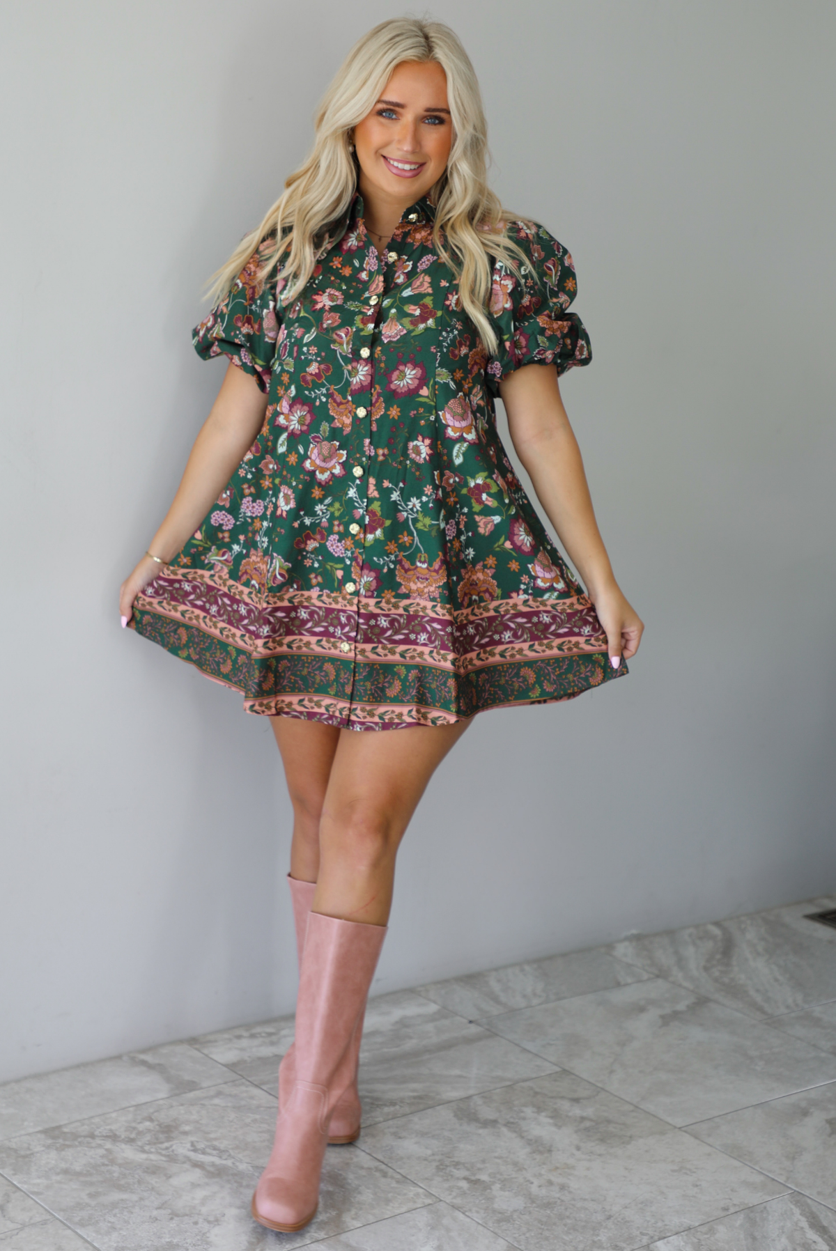 A woman wearing a dark green dress that has a mauve and multicolored floral print pattern throughout, has puffed sleeves, a collared neckline with functional gold button closures in the front, paired with muted pink boots, standing indoors against a white wall with white marble flooring. 