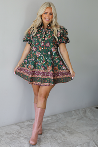 A woman wearing a dark green dress that has a mauve and multicolored floral print pattern throughout, has puffed sleeves, a collared neckline with functional gold button closures in the front, paired with muted pink boots, standing indoors against a white wall with white marble flooring. 