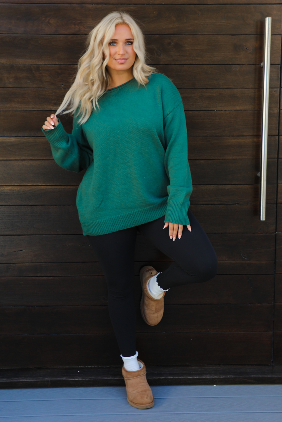 Woman model wearing a green oversized sweater with black leggings, white socks, and tan Uggs. The photo is taken outdoors against a dark brown door. 