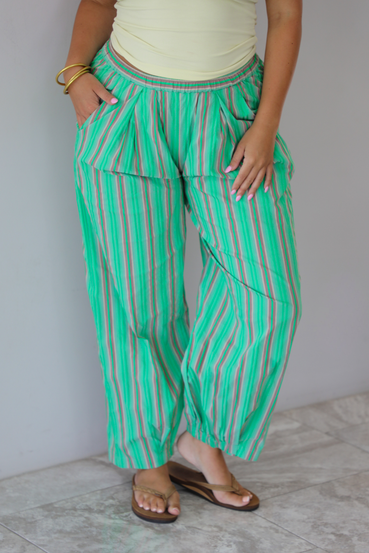 A woman wearing a pale yellow tank and mid-rise pants that have a green, brown, lime green, and tan striped pattern, which have an elastic waistline, functional pockets on either side, and cinched details at the ankles, standing indoors against a white wall with white marble flooring.  The photo is close up and focused on the pants. 