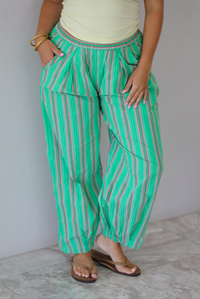 A woman wearing a pale yellow tank and mid-rise pants that have a green, brown, lime green, and tan striped pattern, which have an elastic waistline, functional pockets on either side, and cinched details at the ankles, standing indoors against a white wall with white marble flooring.  The photo is close up and focused on the pants. 