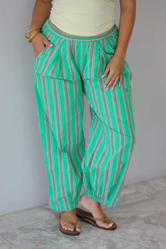 A woman wearing a pale yellow tank and mid-rise pants that have a green, brown, lime green, and tan striped pattern, which have an elastic waistline, functional pockets on either side, and cinched details at the ankles, standing indoors against a white wall with white marble flooring.  The photo is close up and focused on the pants. 