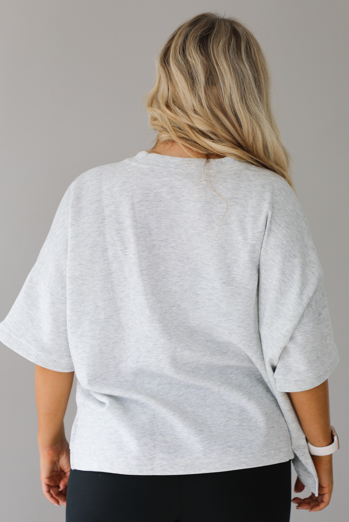 Woman wearing an oversized heather grey box tee with black leggings that flare slightly at the hems. The photo shows a closeup of the back of the tee. The photo is taken indoors against a white wall.