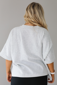 Woman wearing an oversized heather grey box tee with black leggings that flare slightly at the hems. The photo shows a closeup of the back of the tee. The photo is taken indoors against a white wall.