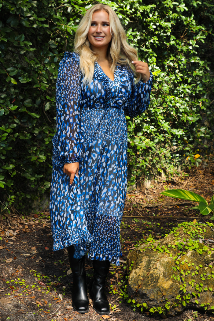 Woman wearing a blue patterned dress and black boots outdoors. The dress has a v-neckline with ruffled details on the bodice, long sleeves, a cinched waistline, with a midi length skirt. 
