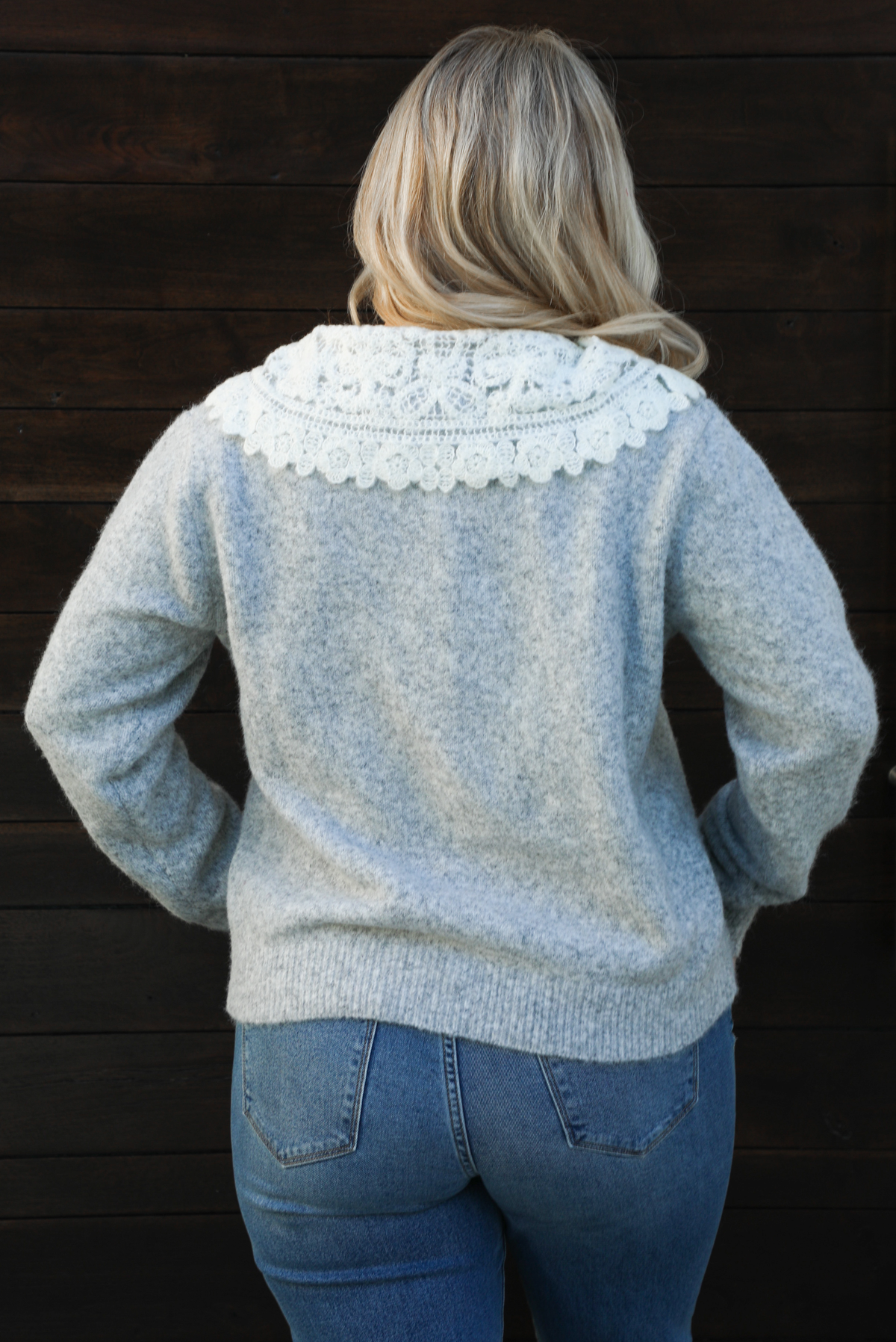 Close up of the back of a woman model wearing a heather grey cardigan with a white lace bib at the neckline, has functional button closures in the front, functional pockets in the front, and a relaxed fit. The cardigan is paired with a medium wash denim and tan ugg boots. The photo is taken outdoors against a dark brown wooden door.