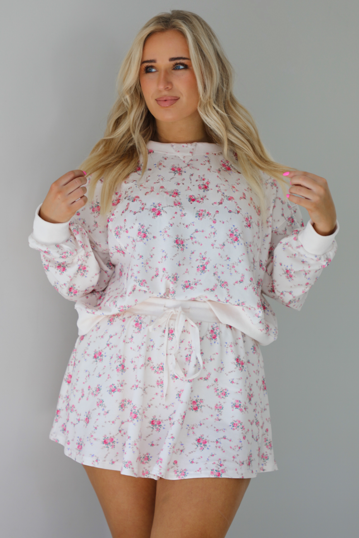 Woman wearing a pale pink two piece set that has a pink floral print pattern throughout. The set featuring an oversized long sleeved top and high waisted skirt. The photo is taken indoors against a white wall.