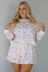 Woman wearing a pale pink two piece set that has a pink floral print pattern throughout. The set featuring an oversized long sleeved top and high waisted skirt. The photo is taken indoors against a white wall.