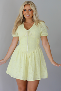 Woman wearing a light yellow mini dress with a floral eyelet design throughout, v-neckline, short sleeves, fitted drop waist bodice, and a flowy mini length skirt indoors against a white wall.