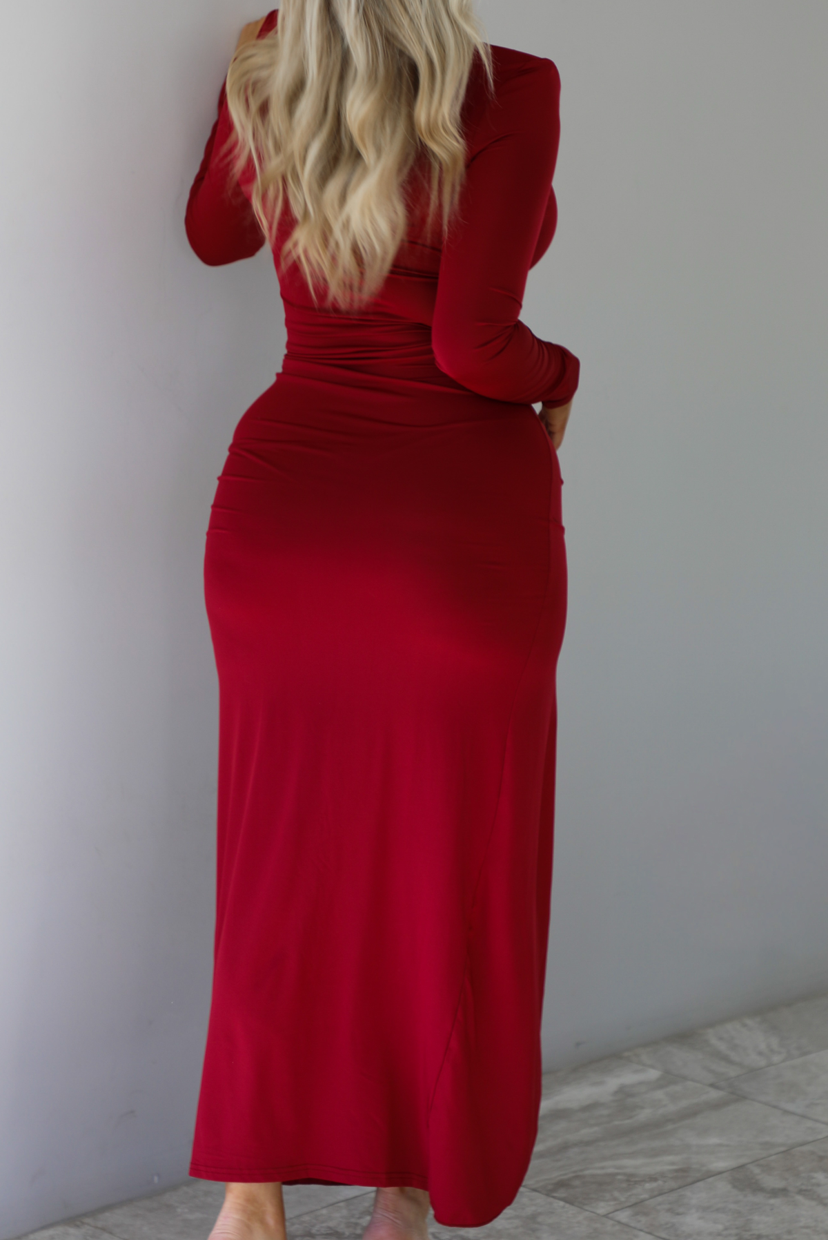 Woman wearing a red maxi dress that has a high neckline, shoulder pads, a fitted bodice, long sleeves, with a split hem detail on the left side. The photo is taken indoors against a white wall with white marble flooring. The photo shows the back of the dress.