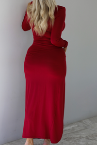 Woman wearing a red maxi dress that has a high neckline, shoulder pads, a fitted bodice, long sleeves, with a split hem detail on the left side. The photo is taken indoors against a white wall with white marble flooring. The photo shows the back of the dress.