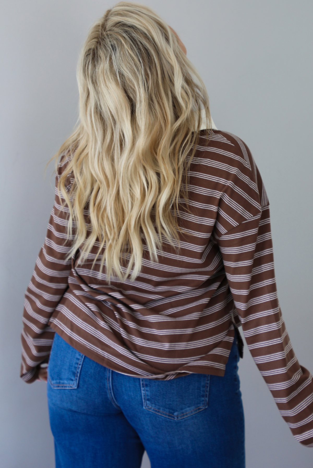 A woman shown from behind wearing a brown and cream striped collared quarter-button long sleeved top, untucked to show the full length, paired with dark denim jeans, standing indoors against a white wall.