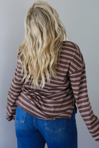 A woman shown from behind wearing a brown and cream striped collared quarter-button long sleeved top, untucked to show the full length, paired with dark denim jeans, standing indoors against a white wall.