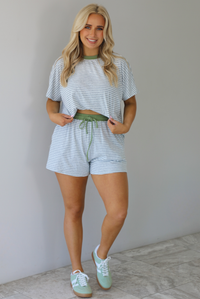 Woman wearing a heather grey and white striped two piece oversized tee and shorts set with green trim at the top of the neckline and on the waistline. The photo is taken indoors against a white wall with white marble flooring. 