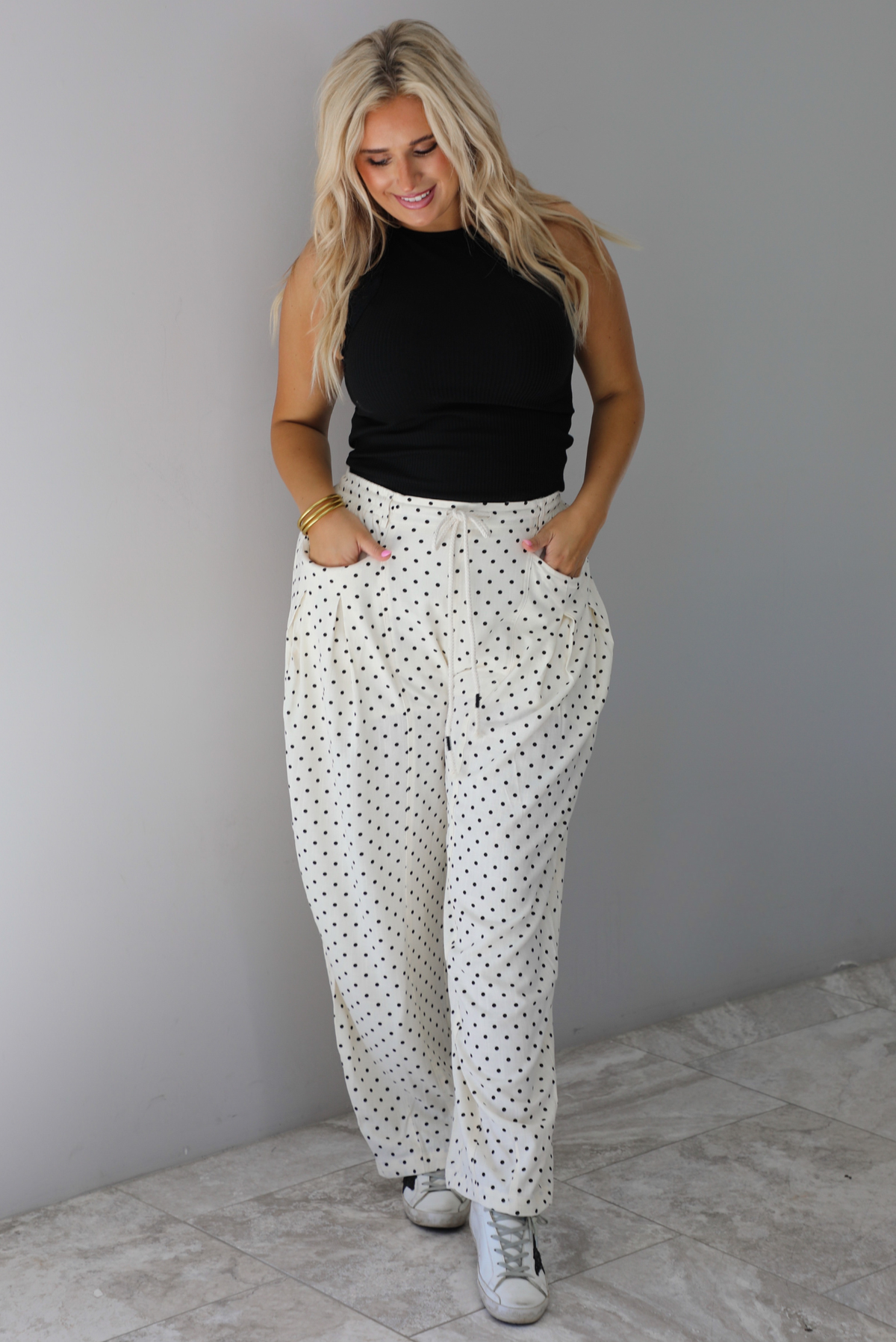 A woman wearing a black high neck tank paired with sand white pants with a black polka dot print pattern throughout, standing indoors against a white wall with white marble flooring. 