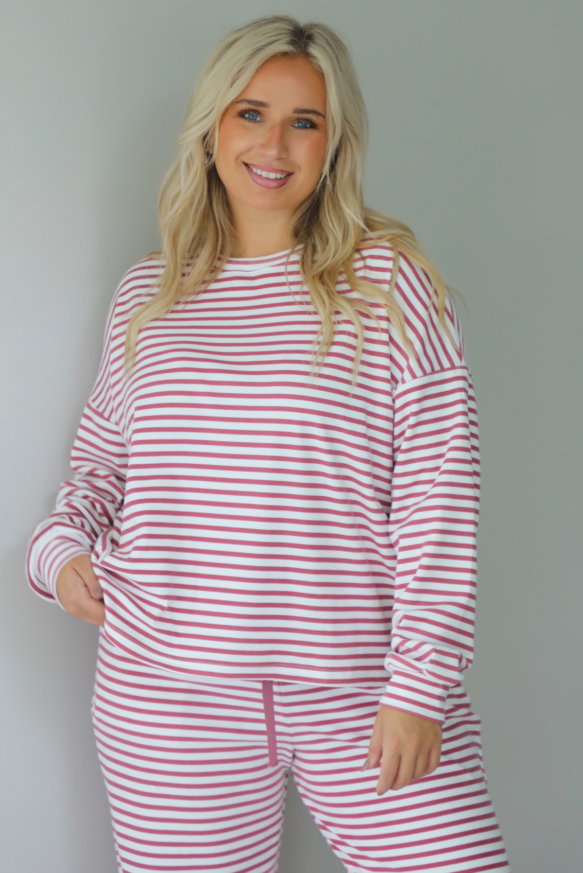 A woman wearing a matching two piece set featuring a long sleeved top and high waisted wide leg pants with a berry and white striped pattern throughout, standing indoors against a white wall. The photo shows the top untucked. 