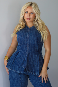 A woman wearing a matching two piece dark wash denim set with a pin striped design on it featuring a sleeveless peplum style top and high waisted denim jeans, standing indoors against a white wall.