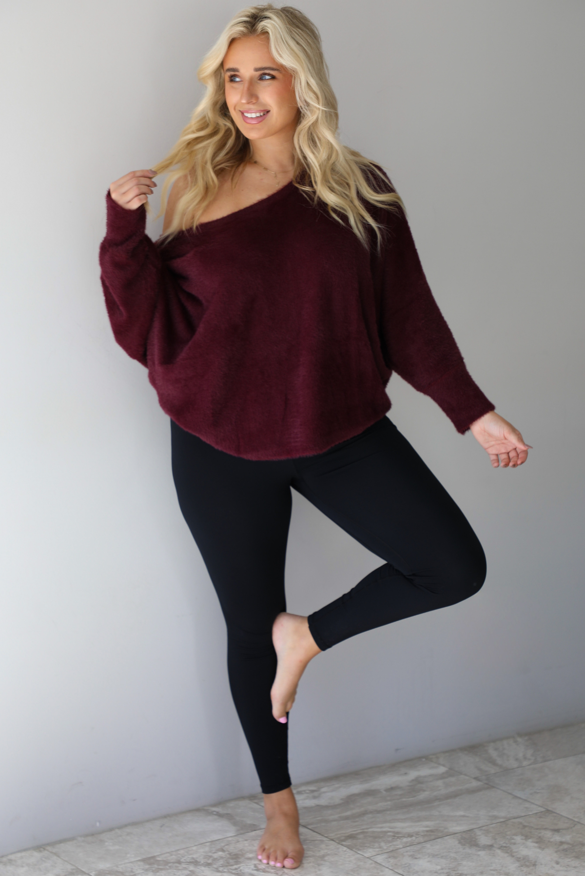 A woman wearing a maroon oversized long sleeved sweater, worn off the shoulder, paired with black leggings, indoors against a white wall with white marble flooring.