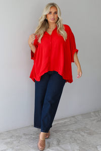 A woman wearing a bright red oversized short-sleeve blouse and dark wide-leg jeans, standing indoors against a white wall with white marble flooring. 
