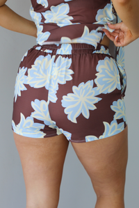 Woman wearing a brown, blue, and off white floral two piece set featuring a vest style top with functional button closures in the front and shorts. The photo is taken in front of a white wall and shows the back of the shorts. 