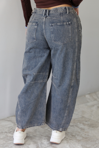 A closeup of a woman shown from behind wearing dark, acid washed barrel denim that have functional back pockets with a long sleeved brown with blue trim cropped top, standing indoors against a white wall with white marble flooring. 