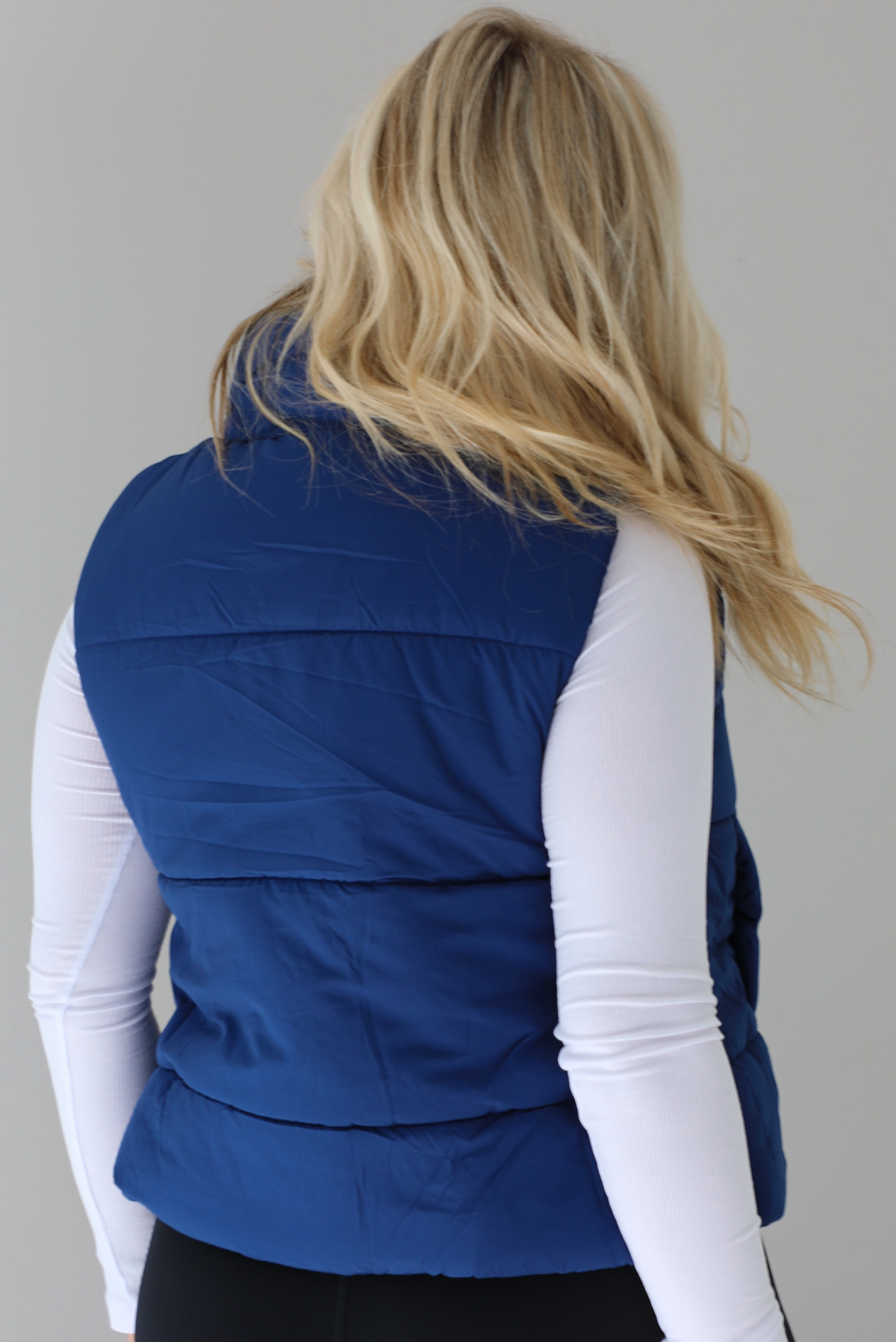 Woman wearing a navy puffer vest, white long-sleeve shirt, and black leggings indoors against a white wall. The puffer vest has a collared design, functional button closures at the neckline, a zipped zipper closure in the front, and functional pockets on either side. The photo shows the back of the vest. 