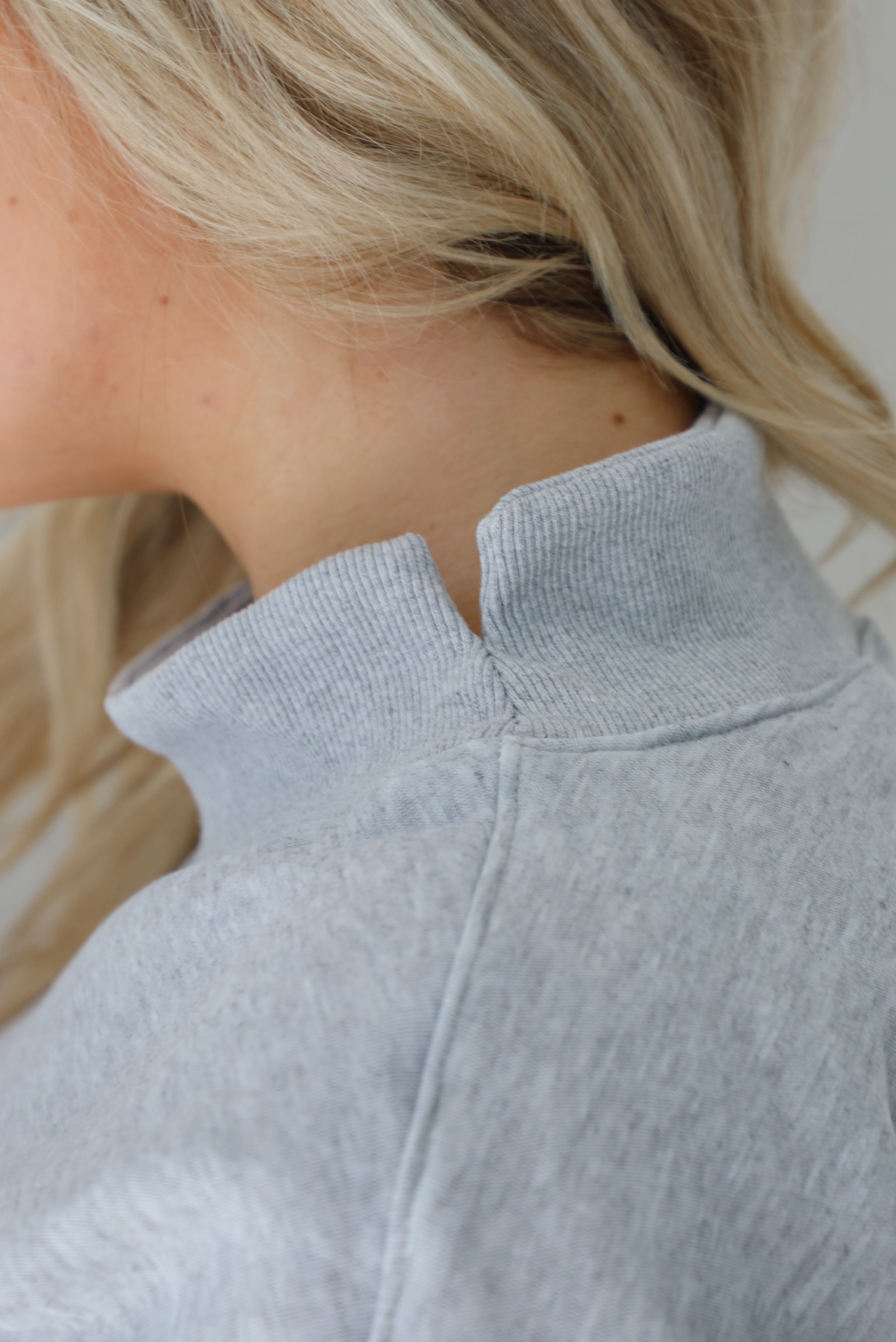 Woman wearing a grey turtleneck sweater, untucked, with high waisted black leggings. The photo shows the split detail at the neckline on the sweater. The photo is taken indoors against a white wall.
