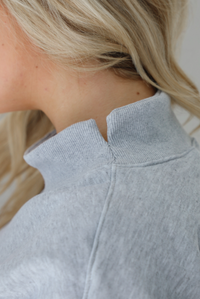 Woman wearing a grey turtleneck sweater, untucked, with high waisted black leggings. The photo shows the split detail at the neckline on the sweater. The photo is taken indoors against a white wall.