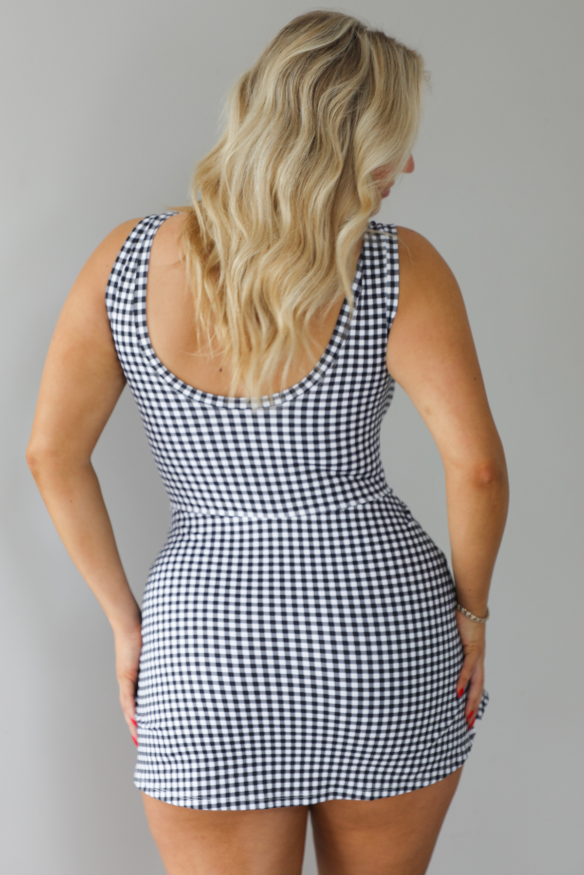 Woman wearing a black and white gingham print athletic romper that has a wrap-style skirt detail. The photo shows the back of the romper that has a scoop back design and the appearance of a dress. The photo is taken indoors against a white wall.