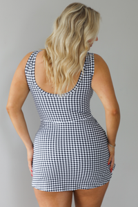 Woman wearing a black and white gingham print athletic romper that has a wrap-style skirt detail. The photo shows the back of the romper that has a scoop back design and the appearance of a dress. The photo is taken indoors against a white wall.