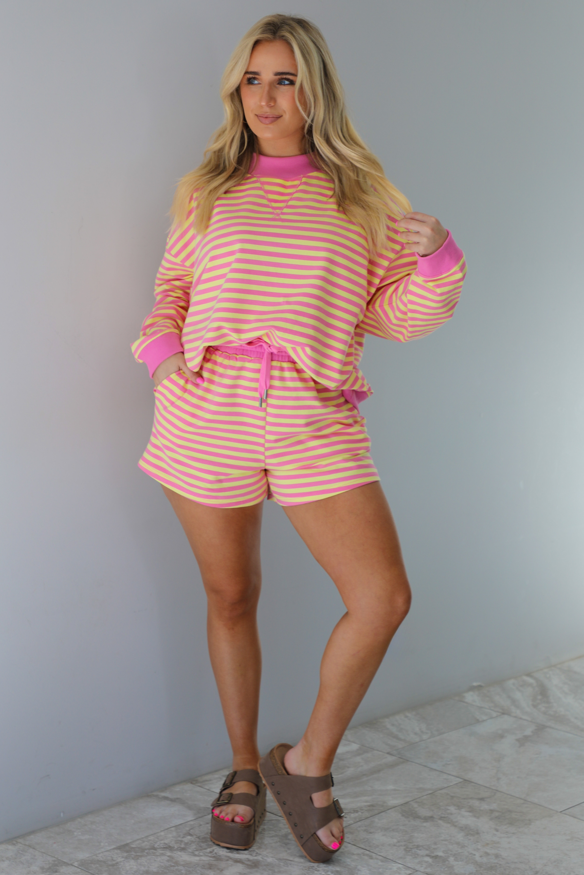 Woman wearing a pink and yellow striped set featuring an oversized sweatshirt and shorts. The photo is taken indoors against a white wall with white marble flooring. 