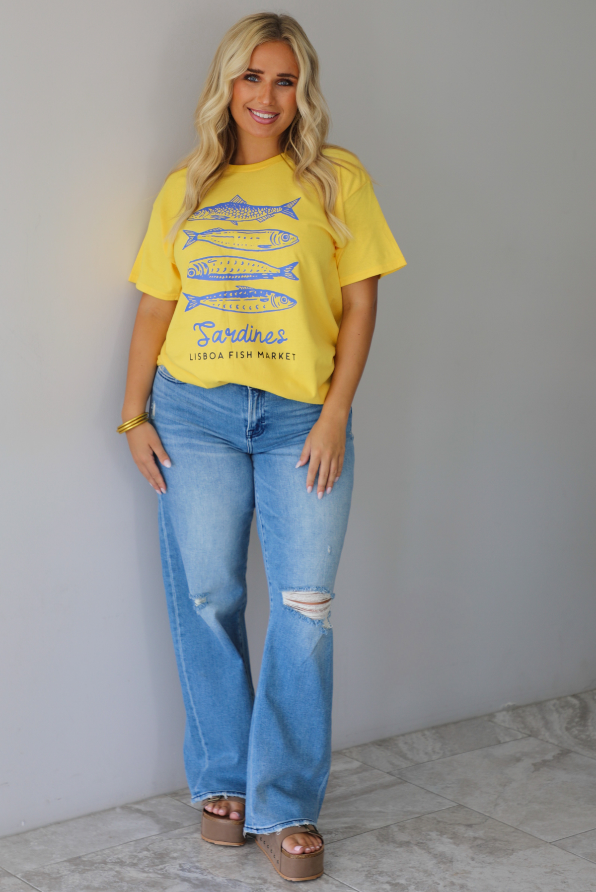 A woman wearing a bright yellow graphic t-shirt with blue sardine illustrations and the text 'Sardines LISBON FISH MARKET', paired with blue jeans, standing indoors against a white wall with white marble flooring. 