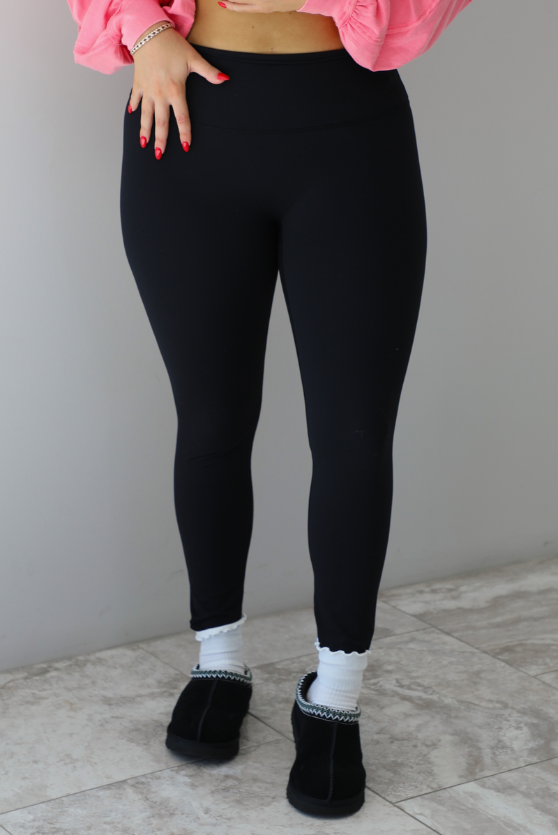 Woman model wearing a fitted pair of black leggings that are high waisted, soft, and super stretchy. She's wearing them with a pair of white ruffled socks and black slip on ugg boot shoes. 